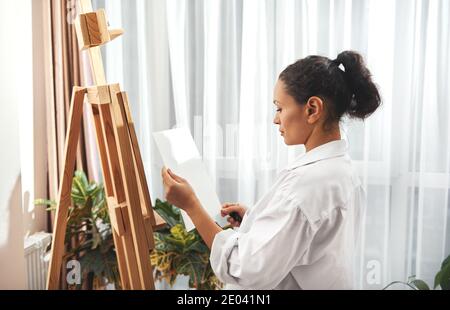Der Künstler hält ein Blatt Aquarellpapier und steht Vor dem Hintergrund einer Staffelei Stockfoto
