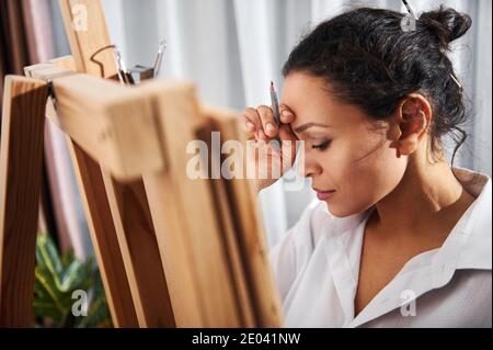 Die junge Künstlerin hält einen Bleistift in den Händen, schaut nach unten und steht vor der Staffelei. Stockfoto