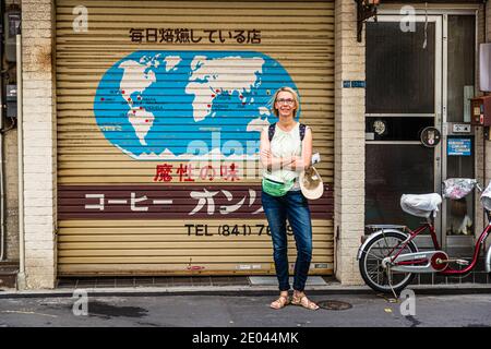 Eine bemalte Weltkarte der Herkunftsländer des Kaffees schmückt das heruntergelassene Rollladen der Kaffeerösterei „Coffee Only“ in der Kappabashi Hon-dori, Tokyos Küchenwarendistrikt im Bezirk Taito, Japan. Angela Berg steht mit verschränkten Armen davor. Der Laden wirbt mit dem Slogan „ein Hauch von Versuchung“ und einem Hinweis, dass die Bohnen täglich geröstet werden. Stockfoto