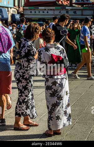 Zwei Frauen in gemusterten Yukatas posieren mit einem Selfie-Stick für ein Foto auf einer belebten Einkaufsstraße in Asakusa, Tokio, Japan. Eine trägt ein rot-schwarzes Blumenmuster, die andere ein weiß-grünes Ahornblattmuster mit einem roten Obi. Stockfoto