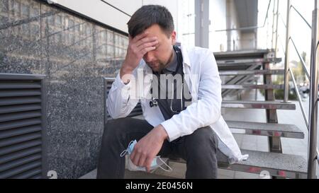 Kaukasischer junger Arzt sitzt auf der Treppe in der Nähe des Klinikgebäudes, müde und unglücklich reiben seine Nase und Augen, Gefühl müde und Kopfschmerzen. Stockfoto