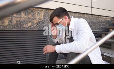 Ernsthafter Arzt, der nach einer sehr langen Schicht im Krankenhaus auf Treppen ruht. Müder Arzt in Maske, weiße Uniform und Stethoskop ruhen, während sie auf der Treppe sitzen Stockfoto