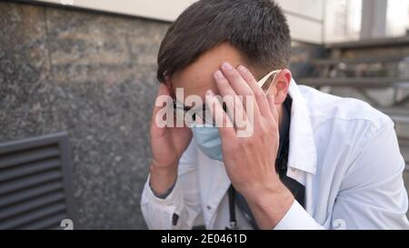 Ernsthafter Arzt, der nach einer sehr langen Schicht im Krankenhaus auf Treppen ruht. Müder Arzt in Maske, weiße Uniform und Stethoskop ruhen, während sie auf der Treppe sitzen Stockfoto