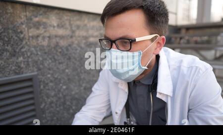 Kaukasischer junger Arzt sitzt auf der Treppe in der Nähe des Klinikgebäudes, müde und unglücklich reiben seine Nase und Augen, Gefühl müde und Kopfschmerzen. Stockfoto