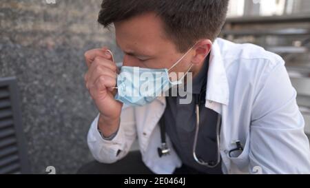 Ernsthafter Arzt, der nach einer sehr langen Schicht im Krankenhaus auf Treppen ruht. Müder Arzt in Maske, weiße Uniform und Stethoskop ruhen, während sie auf der Treppe sitzen Stockfoto