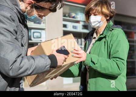 Lieferung Mann trägt eine Gesichtsmaske und ein Paket an die Frau mit einer Maske geliefert, halten Sie die soziale Distanz Stockfoto