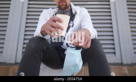 Hände Nahaufnahme mit Tasse Kaffee und schützende medizinische Maske entspannen nach der Erholung des Patienten und harte Arbeit. Gesundheitsarbeiter fühlt sich nach dem Schutz müde Stockfoto