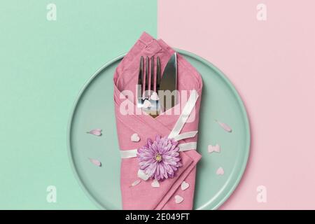 Schöne festliche Tischdekoration mit grünem Teller, Serviette, Blumen und Herzen auf pastellrosa und grünem Hintergrund. Draufsicht mit Kopierbereich. Stockfoto