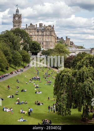 Menschen, die die Sommersonne in den Princes Street Gardens im Zentrum von Edinburgh genießen, mit dem Balmoral Hotel im Hintergrund. Stockfoto