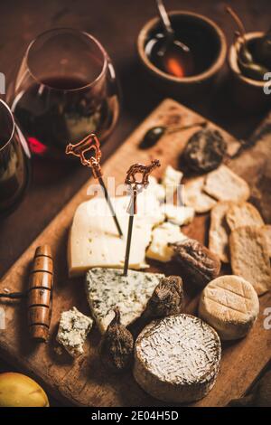 Auswahl an Snacks und Vorspeisen für Rotwein. Verschiedene Käsesorten auf Stöcken mit Stier, Cracker, Trockenobst und Gläser Rotwein auf rustikalem Holzbrett über rostbraunem Hintergrund, selektiver Fokus Stockfoto