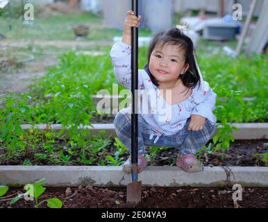 Ein süßes asiatisches Mädchen hilft ihrer Familie, Gemüse in ihrem Hinterhof anzubauen, mit einem Spaten, um den Boden auszugraben und für die Aussaat vorzubereiten. Stockfoto