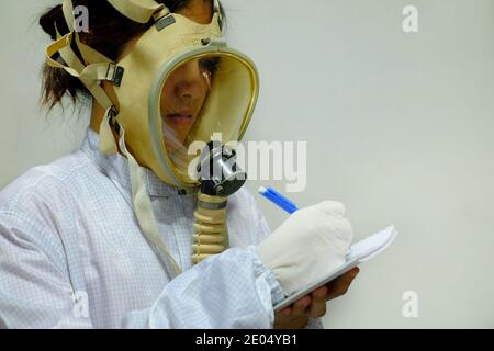 Eine Frau mit einem vollen Gesicht schützende Sauerstoffmaske, weißem Mantel und Gummihandschuhe, immer bereit, in einem biochemischen Labor zu arbeiten, Notizen mit ihrem Stift A Stockfoto