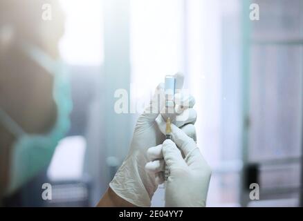 Eine asiatische Ärztin mit weißen Gummihandschuhen und einer OP-Maske in einer Klinik, die eine Nadel und eine Spritze auf eine klare Glasflasche mit Impfstoff einführt Stockfoto