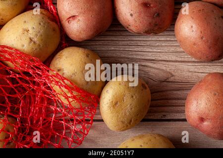 Flat Lay Nahaufnahme Bild mit einem roten Netz Kartoffelsack mit rosa und gelben rohen Bio-Kartoffeln auf Holzhintergrund. Ein vielseitiges Stillleben Essen Stockfoto