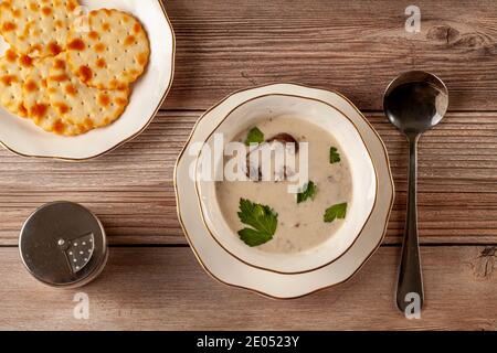 Flaches Lay-Bild von cremigen Pilzsuppe mit Petersilie verziert Blätter in einem handwerklichen Porzellanbecher auf einem flachen Teller Auf einem Holztisch mit Cracker serviert Stockfoto