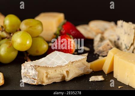 Eine Auswahl an französischen, italienischen und Schweizer Käsesorten in Höhlenalter auf schwarzem Käseplatte, serviert mit Crackern und Früchten (Trauben und Strohhalmchen) Stockfoto