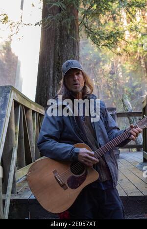 Ein Mann mittleren Alters, der einen Hut trägt, der Gitarre an der Point View Bridge im Park von West Vancouver, BC/Kanada - November 8,2020 spielt. Blick auf die Straße Stockfoto