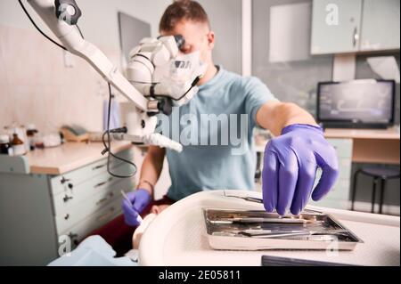 Nahaufnahme der männlichen Zahnarzthand in sterilen Handschuh hält Metall Dental Explorer beim Überprüfen der Zähne des Patienten. Stomatologe nimmt Instrument, während er in der Nähe des Mikroskops sitzt. Konzept der Zahnmedizin. Stockfoto