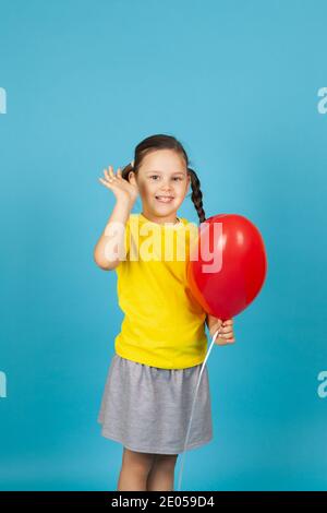 Ein fröhliches, fröhliches Mädchen mit Zöpfen in einem gelben T-Shirt hält einen roten Ballon in Herzform und winkt ihre Hand, isoliert auf blauem Hintergrund Stockfoto