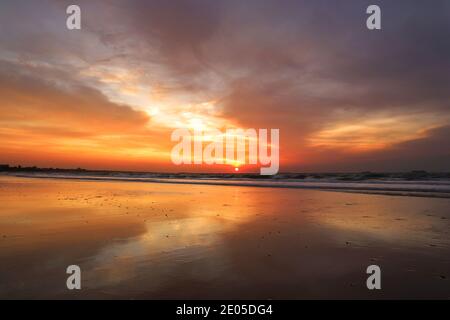 Die Sonne geht in feurigem Orange und Rot in einen wolkigen Winterhimmel über, der sich im nassen Sand des Bournemouth Beach widerspiegelt. Stockfoto