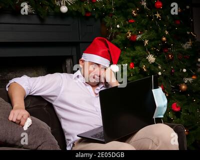 Ein trauriger, kranker Mann in einem Weihnachtsmann-Hut schaut sich allein einen Laptop an. Die Feier des neuen Jahres und die Epidemie. Stockfoto