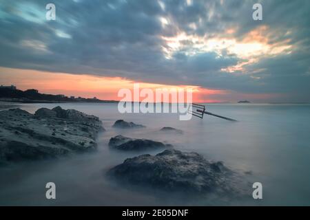 Das Meer wirbelt und stürzt um die Felsen einer Meeresschutzgroyne am Bournemouth Beach, während die aufgehende Sonne durch die Wolkendecke bricht. Stockfoto