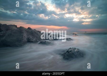 Das Meer wirbelt und stürzt um die Felsen einer Meeresschutzgroyne am Bournemouth Beach, während die aufgehende Sonne durch die Wolkendecke bricht. Stockfoto