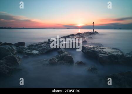 Das Meer wirbelt und stürzt um die Felsen einer Meeresschutzgroyne am Bournemouth Beach, während die aufgehende Sonne durch die Wolkendecke bricht. Stockfoto