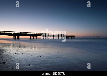 Die Silhouette des Boscombe Pier erstreckt sich am Horizont unter einem kalten, tiefblauen Himmel, wenn die Sonne aufgeht. Stockfoto