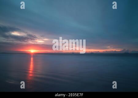 Die aufgehende Sonne gutet durch die Wolkendecke und feuert einen warmen, orangen Lichtwellenzug über den nassen Sand des Bournemouth Beach. Stockfoto