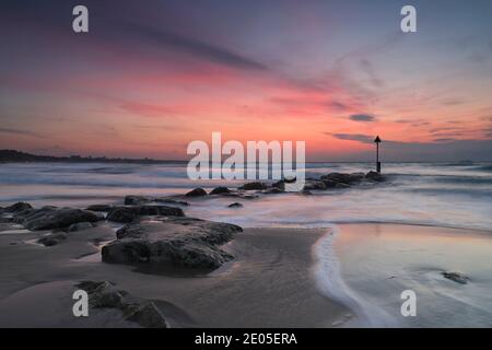 Wispy Wolken spiegeln den Verlauf eines felsigen Meeresschutzrauschens wider, der zur aufgehenden Wintersonne führt, während die Wellen auf den Bournemouth Strand Rollen. Stockfoto