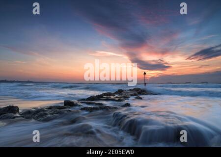 Die Wellen fallen über die großen Felsen eines Meeresschutzrauschens, während die Sonne am Bournemouth Beach in einen kalten Winterhimmel voller wispiger Wolken aufgeht. Stockfoto