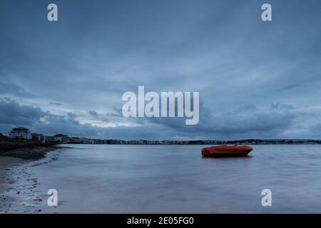 Ein kleines orangefarbenes Boot thront auf sanften Wellen, während düstere graue Wolken über der sich verjüngenden Skyline von Sandbanks schwer am Himmel hängen. Stockfoto