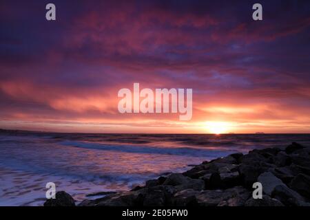 Die aufgehende Sonne füllt einen Himmel voller traumhafter, wispiger Wolken mit magischen rosa und violetten Farben, während die Wellen in schaumhaftem Chaos auf das Ufer stürzen. Stockfoto