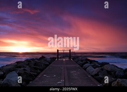 Die aufgehende Sonne füllt einen Himmel voller traumhafter, wispiger Wolken mit magischen rosa und violetten Farben über den Führungslinien einer Meeresverteidigungsgroyne. Stockfoto