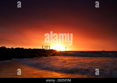 Das goldene Licht der aufgehenden Sonne erhellt die dicken Winterwolken und füllt sie mit feurigen Rottönen, während eine Welle zum Ufer hinaufeilt. Stockfoto