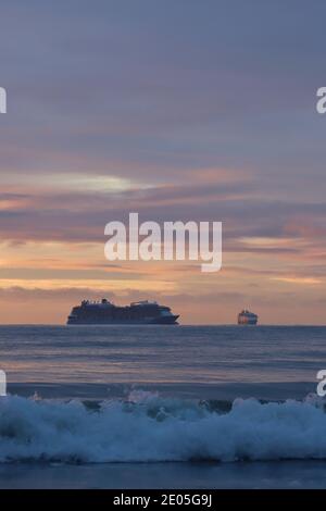 Ein Paar Kreuzschiffe kann am Horizont unter einem Himmel voller pastellfarbener Wolken gesehen werden, während die Wellen in einem weißen Schaum zum Ufer Rollen. Stockfoto