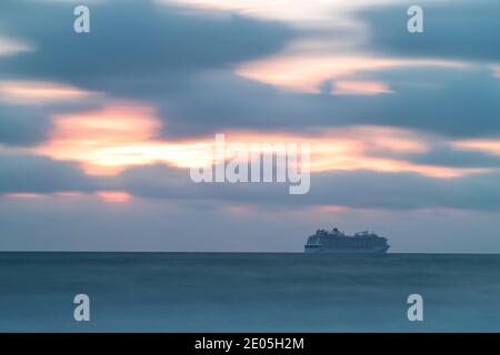 Ein riesiges Kreuzschiff geht am Horizont entlang, während die aufgehende Sonne goldenes Licht zwischen dicken grauen Wolken in den Winterhimmel feuert. Wolken ziehen über dem Himmel. Stockfoto