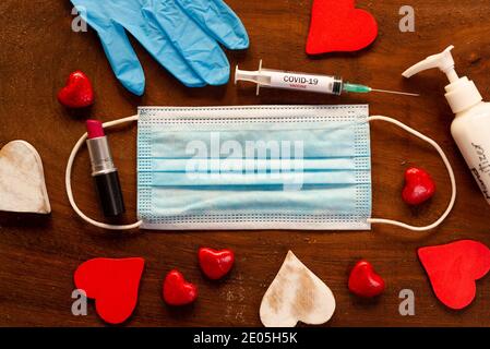 Covid valentinstag Hygiene-Konzept. Gesichtsmaske mit roten Herzen auf Holz Hintergrund. Draufsicht der Hand Desinfektionsmittel und Lippenstift und Spritze für den Urlaub