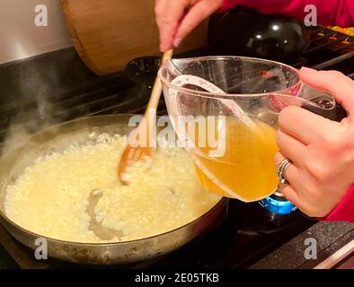 Eine Hauskochin gießt Brühe in ihr Reis Risotto unter Rühren in einer Pfanne auf einem Gasherd. Stockfoto