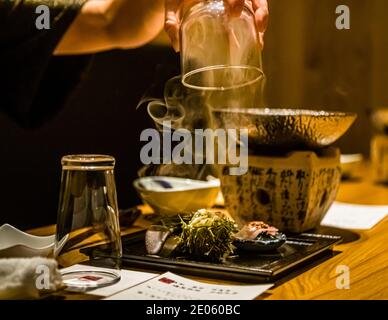 Ein Kellner hebt ein Glas von einem dampfenden Fischgericht. Daneben ein kunstvoll arrangierter Beilagensalat aus Kräutern und Seetang. Restaurant in Shizuoka, Präfektur Shizuoka, Japan. Stockfoto