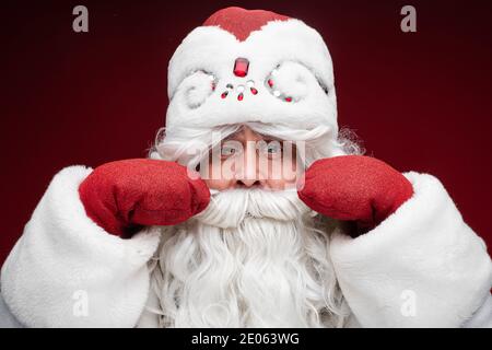 Älterer grauhaariger Schnurrbart bärtiger Weihnachtsmann in Weihnachtsmütze auf rotem Studioportrait. Weihnachten Neujahr 2021 Feier Stockfoto
