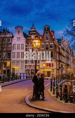 Amsterdam Niederlande bei Sonnenuntergang, historische Kanäle bei Sonnenuntergang. Holländische historische Grachten in Amsterdam, Paar auf Städtereise in Amsterdam, Männer und Frauen am Abend an den Grachten Stockfoto