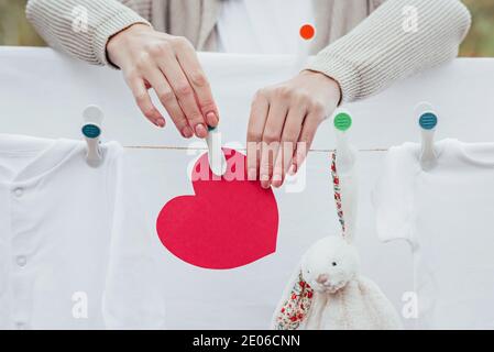 Die Hände der Frau hängen an der Wäscheleine gewaschene Babykleidung und rotes Herz mit Plastikstiften. Selektiver Fokus, Außenansicht. Stockfoto