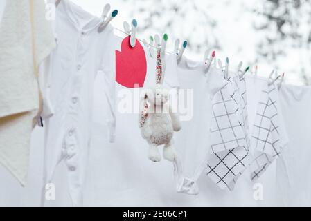 Weiß gewaschene Babykleidung, Spielzeug und rotes Herz auf Wäscheleine mit Plastikstiften. Selektiver Fokus, Außenansicht. Stockfoto