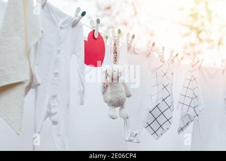 Weiß gewaschene Babykleidung, Spielzeug und rotes Herz auf Wäscheleine mit Plastikstiften. Selektiver Fokus, Außenansicht. Stockfoto