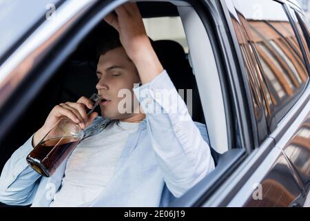 Betrunkener, schläfriger Mann, der im Auto sitzt und mit geschlossenen Augen Whiskey trinkt Stockfoto