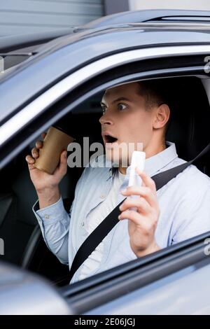 Schockiert Mann hält Kaffee zu gehen und Handy auf Fahrer Sitz im Auto, verschwommen Vordergrund Stockfoto