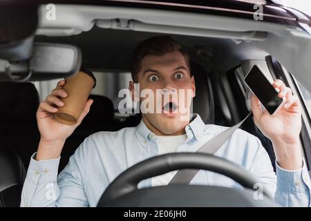 Erschrocken Mann hält Kaffee zu gehen und Smartphone am Lenkrad im Auto, verschwommen Vordergrund Stockfoto