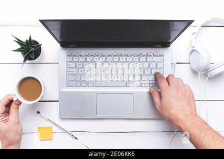 Flach legen, Hände klopfen eine Laptop-Tastatur und halten eine Tasse Kaffee auf weißem Holztisch. Stockfoto